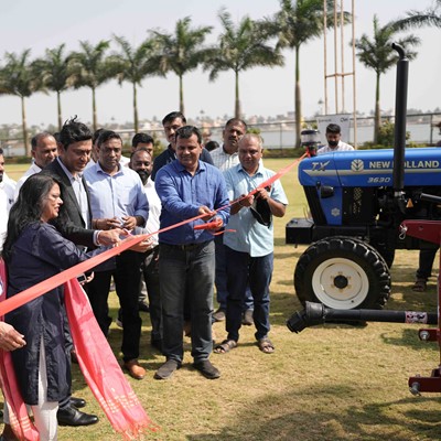 A group of people gathered outdoors on a grassy area with palm trees in the background, participating in a ribbon-cutting ceremony. A woman and a man in the center are cutting a red ribbon with scissors, while others stand around them watching. A blue New Holland tractor is visible on the right side of the image. The event appears to be a formal or celebratory occasion.