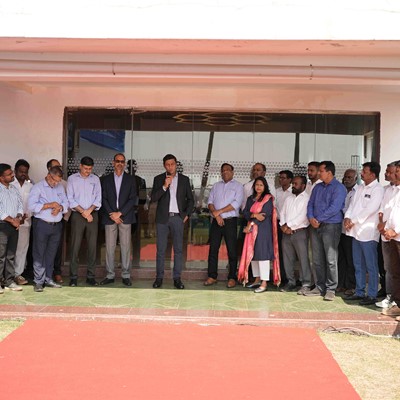 A group of professionally dressed men and women standing in a semi-circle outside a building with a red carpet laid out in front. One man in the center is speaking into a microphone, suggesting a formal event or gathering. The background shows glass doors and a white exterior wall.