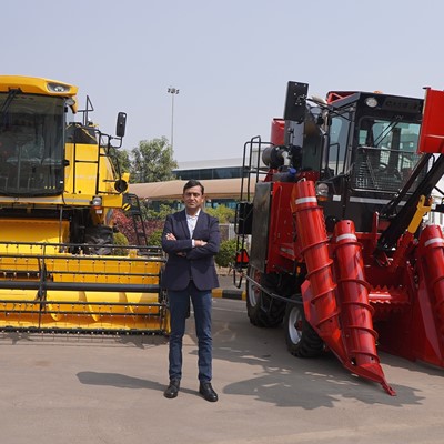 A man in a dark blazer and jeans stands with arms crossed between two large agricultural machines, one yellow and one red, parked on a paved area outside a building under a clear sky.