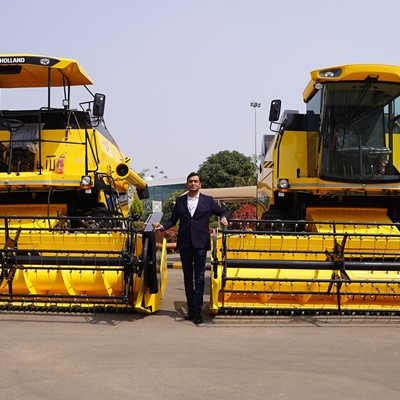 A man in a dark suit standing between two large yellow New Holland agricultural combine harvesters parked outdoors on a paved surface, with a building and greenery in the background under a clear sky.