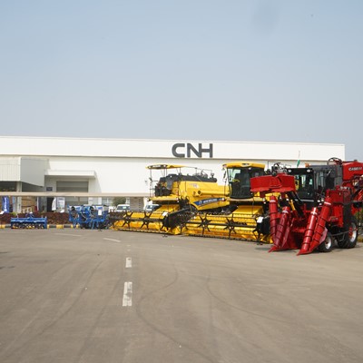 Outdoor display of large agricultural machinery including yellow combine harvesters and a red corn harvester parked in front of a white industrial building with the CNH logo, under a clear blue sky.
