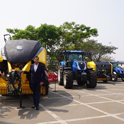 A man in a dark suit stands next to large yellow and black agricultural machinery and blue tractors parked in a row on a paved outdoor area with trees and red barriers in the background under a clear sky.