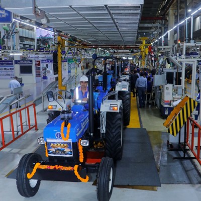 A blue tractor decorated with orange marigold garlands is being driven by a worker wearing a white helmet and reflective vest inside a large, well-lit industrial factory. The factory floor is busy with machinery, equipment, and other workers in the background, indicating a manufacturing or assembly line environment.