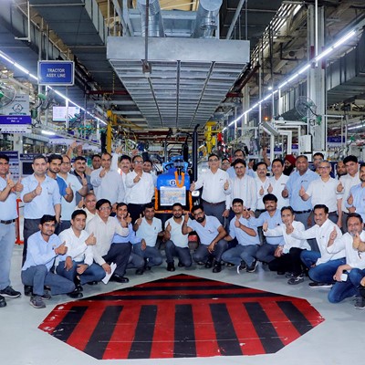 A large group of factory workers and engineers posing together inside a manufacturing facility, all giving thumbs-up gestures. They are standing and squatting around a blue tractor assembly line area, with industrial equipment and machinery visible in the background. The environment is well-lit with overhead lights and organized workstations.