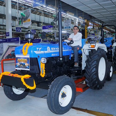 A man sitting on a blue New Holland TX 3600-2 tractor decorated with orange marigold garlands inside a factory assembly line. The tractor has a sign indicating "800000" units produced. Several similar tractors are lined up behind it, and factory workers are visible in the background. The setting is a well-lit industrial environment with various signs and equipment.