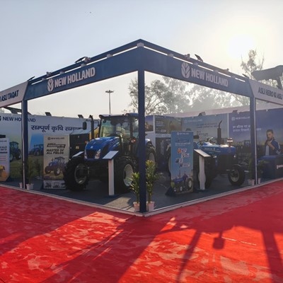 Outdoor exhibition booth of New Holland showcasing blue tractors on a red carpeted area with banners and informational displays, under a clear sky with sunlight and trees in the background.