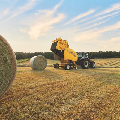 A blue tractor with a yellow hay baler attachment is working in a large open field during sunset, creating round hay bales. One large hay bale is in the foreground on the left, with more hay bales scattered across the field and a line of trees in the background under a partly cloudy sky.