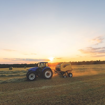 A blue tractor pulling a yellow baler machine in a large open field during sunset, with round hay bales scattered across the field and a clear sky with soft clouds in the background.