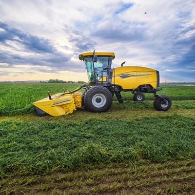 A yellow New Holland agricultural harvester machine is parked on a green field under a partly cloudy sky during daylight, with freshly cut crops visible on the ground.
