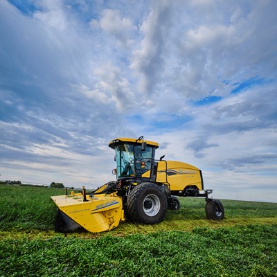 A yellow agricultural mower machine cutting green grass in a vast field under a partly cloudy blue sky during daytime.