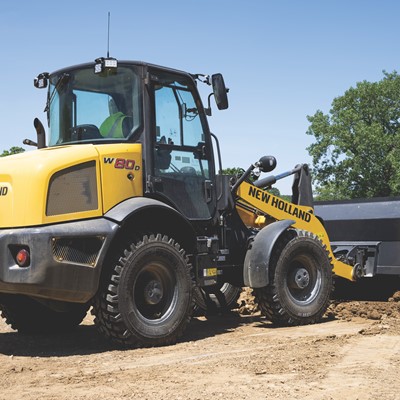 Yellow New Holland W80D wheel loader with a front bucket filled with dirt, parked on a construction site with trees and a clear blue sky in the background.