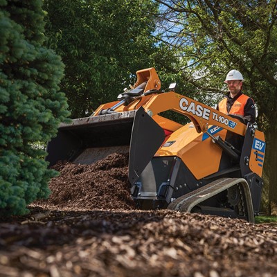 A worker wearing a white hard hat and orange safety vest operates a yellow CASE TL100EV electric mini track loader spreading mulch around a landscaped area with trees and greenery in the background