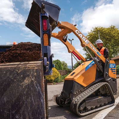 A worker wearing a white hard hat and orange safety vest operates a yellow CASE TL100EV electric mini track loader dumping mulch into a large container on a sunny day with blue sky and scattered clouds
