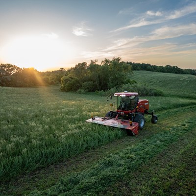 A red agricultural mower cutting grass in a large green field during sunset, with trees and rolling hills in the background under a partly cloudy sky.