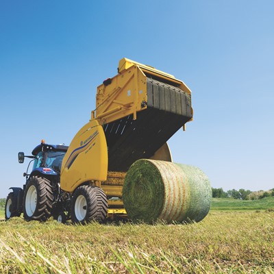 A blue tractor attached to a yellow New Holland round baler machine is operating in a green field under a clear blue sky, producing a large round bale of hay. In the background, there are some trees and a red barn.