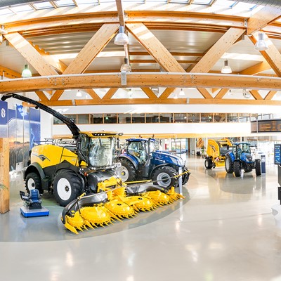 Interno di un moderno showroom agricolo con macchinari agricoli di grandi dimensioni, tra cui trattori blu, mietitrebbie gialle e attrezzature per la raccolta, illuminato da ampie finestre e travi in legno a vista.