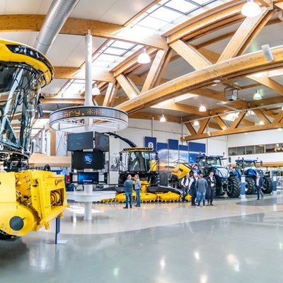Interno di un grande showroom con macchinari agricoli moderni di colore giallo e blu, tra cui mietitrebbie e trattori, con alcune persone che osservano e discutono vicino ai veicoli, struttura con travi in legno e ampie finestre che lasciano entrare molta luce naturale.