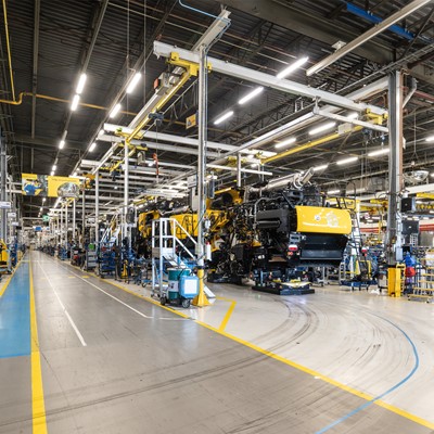 Interno di una grande fabbrica moderna con linee di assemblaggio di macchinari pesanti gialli, illuminazione a LED e pavimenti segnati per la sicurezza, senza operai visibili.