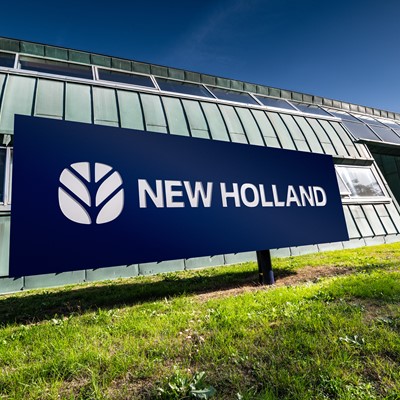 Insegna blu con il logo e la scritta "NEW HOLLAND" davanti a un edificio moderno con pareti in metallo verde e finestre, su un prato verde sotto un cielo sereno.