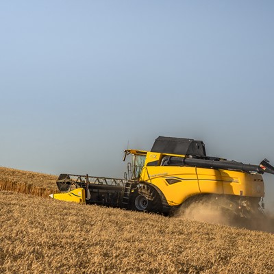 A yellow combine harvester working on a golden wheat field under a clear blue sky, harvesting crops and kicking up dust as it moves along a sloped terrain.