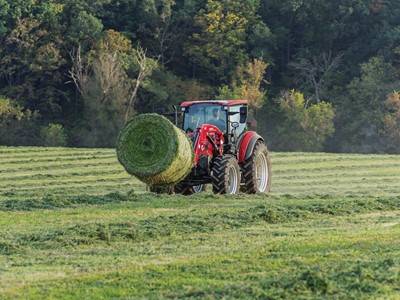 A red tractor pulling a large round hay bale across a lush green field, surrounded by trees and a clear sky. The scene captures the essence of agricultural work and rural life.