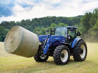 A blue New Holland tractor is lifting a large round hay bale in a grassy field. The background features lush green trees and a cloudy sky, indicating a rural farming setting. The tractor's front loader is engaged, showcasing its capability for handling heavy loads.