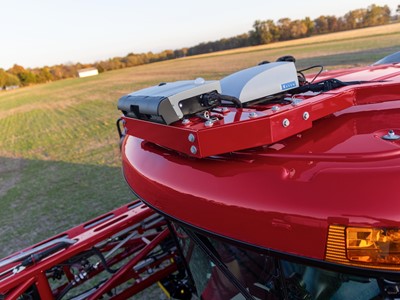 A close-up view of the top of a red agricultural machine, featuring mounted equipment including sensors and communication devices. The background shows a green field under a clear sky, indicating a rural farming environment.