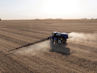A blue tractor equipped with a sprayer is working in a vast, empty agricultural field. Dust is being kicked up as the tractor moves across the dry, brown soil under a clear sky. The landscape is flat and stretches into the distance, showcasing the agricultural setting.