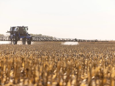 A large agricultural sprayer tractor applying chemicals to a field of dry, golden crops under a clear sky. The tractor is positioned in the distance, with a long spray arm extending outward, creating a mist as it moves through the harvested field.