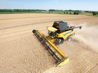 A yellow combine harvester working in a vast field of golden crops under a clear blue sky. Dust is being kicked up as the machine moves through the field, showcasing the agricultural landscape with green trees in the background.