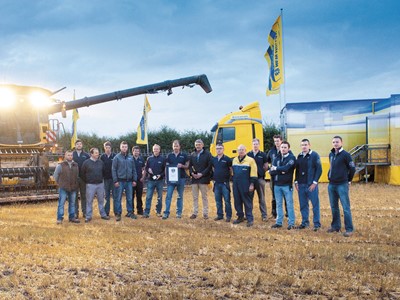 A group of 15 people stands in a field during twilight, with a large agricultural machine and a truck in the background. The individuals are dressed in casual work attire and are smiling, holding a plaque. Yellow flags with a logo are visible, indicating a celebration or event related to farming or agriculture. The setting appears to be a harvest or agricultural demonstration.