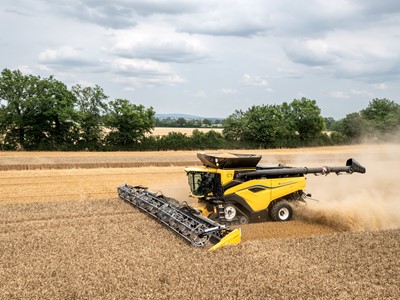 A yellow and black combine harvester is working in a field, harvesting golden wheat. Dust is being kicked up as the machine moves through the crop, with green trees and a cloudy sky in the background. The scene captures the essence of agricultural activity during the harvest season.