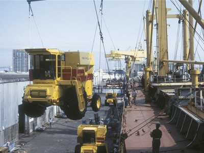 A yellow agricultural harvester being lifted by a crane at a port, with cargo ships and workers in the background. The scene depicts industrial activity, showcasing the transportation of heavy machinery.
