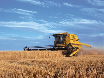 A yellow combine harvester working in a golden wheat field under a blue sky with scattered clouds. The machine is cutting through the tall wheat stalks, creating a cloud of dust.
