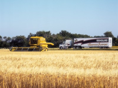 A yellow combine harvester is working in a golden wheat field, with a large truck bearing the "New Holland Harvest Support" logo parked nearby. The background features trees and a clear blue sky, emphasizing a rural agricultural setting.