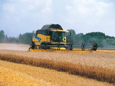 A yellow and black combine harvester working in a golden wheat field under a blue sky with scattered clouds. Dust is being kicked up as the machine moves through the crops, surrounded by green trees in the background.