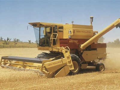 A yellow New Holland combine harvester working in a golden wheat field under a clear blue sky. The machine is actively harvesting crops, with dust rising around it, showcasing agricultural machinery in action.