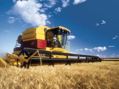 A New Holland combine harvester working in a golden wheat field under a blue sky with scattered clouds. The machine is prominently featured, showcasing its yellow and red colors as it cuts through the tall wheat.