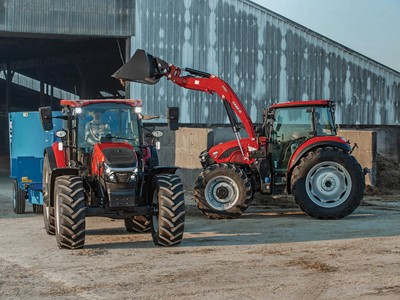 Two red tractors are positioned in front of a large barn with a corrugated metal exterior. One tractor is equipped with a front loader lifting a bucket filled with material, while the other tractor is attached to a blue trailer. The scene appears to be on a farm with dirt ground and hay visible near the barn.