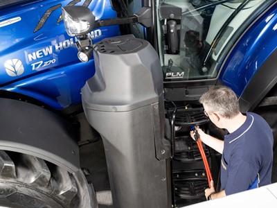 A man in a navy blue shirt is inspecting or repairing the steps of a blue New Holland T7.270 tractor inside a workshop or garage. The tractor's large front tire and side mirror are visible, along with the "Methane Power" label on the tractor's hood.