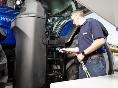 A man wearing a navy blue polo shirt and jeans is inspecting or refueling a large blue New Holland tractor. He is holding a red hose connected to a nozzle near the tractor's steps, standing next to the tractor's large rear tire. The scene appears to be outdoors under a covered area.