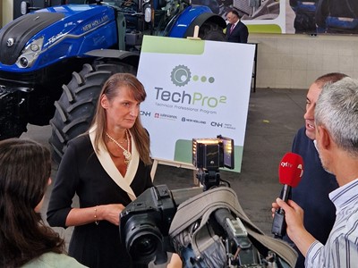 A woman is being interviewed by a news crew inside a workshop or showroom with a blue New Holland tractor in the background. Behind her is a large sign displaying the "TechPro2" logo and the text "Technical Professional Program." The setting includes posters of people working on machinery, emphasizing a technical or agricultural theme. The interviewers hold microphones and a professional video camera is recording the scene.