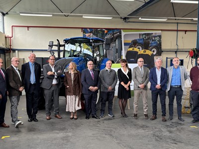 A group of twelve professionally dressed people standing in a row inside an industrial workshop with a large blue tractor behind them. The setting appears to be a formal event or visit, with a mix of men and women posing for a group photo. The background includes industrial equipment, posters, and workshop lighting.