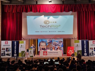 A large audience seated in an auditorium facing a stage with red curtains. On the stage, a speaker stands at a podium with a microphone, presenting in front of a large screen displaying the "TechPro² Technical Training Program" logo. Banners for Salesianos, New Holland, and CNH Industrial flank the stage. The event appears to be a formal presentation or training session.