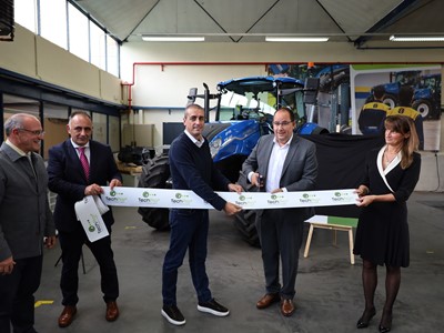 Five people standing in a workshop with a blue tractor in the background, participating in a ribbon-cutting ceremony. One man in a gray suit is cutting a white ribbon with "TechPro" logos, while the others hold the ribbon and smile. The setting appears to be an industrial or agricultural equipment facility.