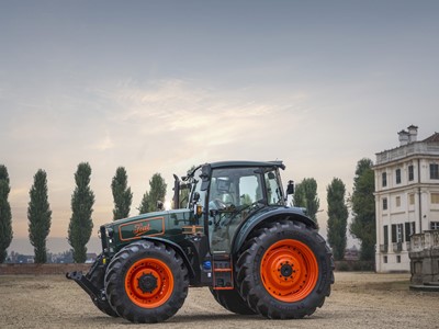 A green and orange Fiat tractor parked on a dirt ground with tall trees in the background and a large white building on the right side under a cloudy sky during sunset.
