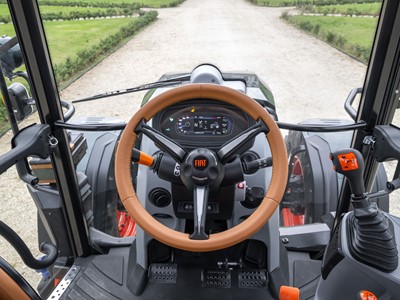 Interior view of a Fiat tractor cabin showing the steering wheel with a brown leather cover, dashboard with digital display, control levers, pedals, and a clear view of the gravel path and green fields outside.