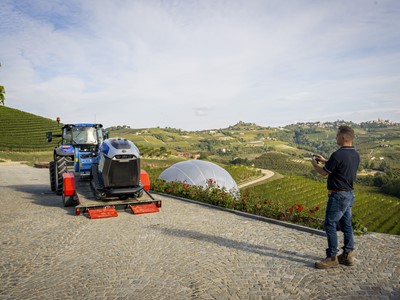 A man operating a remote control stands on a cobblestone path overlooking a scenic vineyard landscape with rolling hills. In front of him, a blue tractor is carrying a modern robotic agricultural machine on a trailer. The vineyard rows stretch across the hills under a partly cloudy sky, with a white dome structure visible among the greenery.