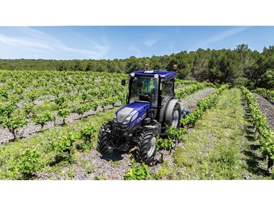 A blue tractor is driving through a vineyard with rows of green grapevines under a clear blue sky, surrounded by a forest in the background.