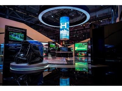 Exhibition booth showcasing advanced agricultural technology with a blue autonomous robotic vehicle on a circular platform, multiple digital screens displaying field preparation and seed planting information, and a large cylindrical hanging digital display with the word "Tech" in a modern indoor event space.